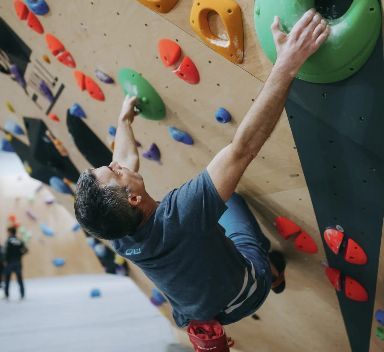 Man climbing an indoor rock wall with colourful holds whilst other climbers train in the background.