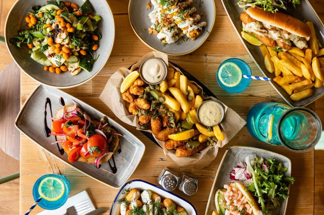 Overhead view of a wooden table spread with various dishes including salads, fish and chips, burgers, and fried appetisers, with drinks and condiments.