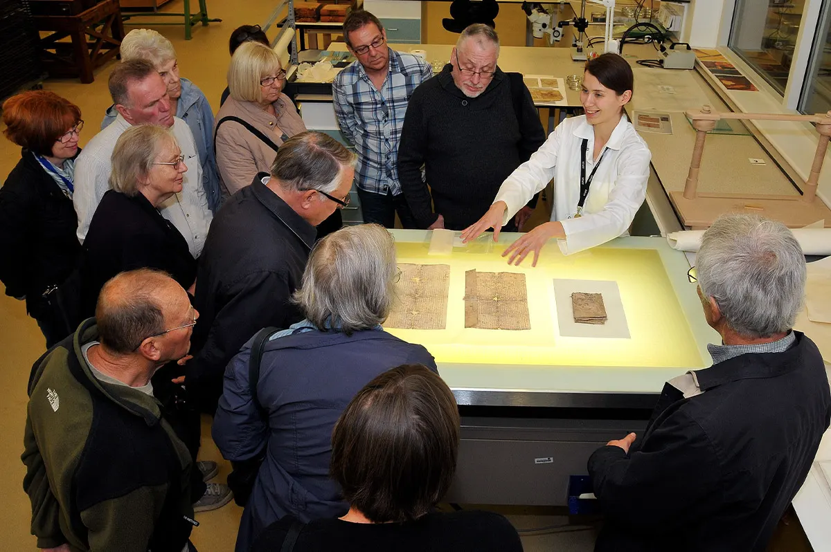 A tour guide in white presents historical documents illuminated on a display table to a group of visitors gathered around in a museum setting.