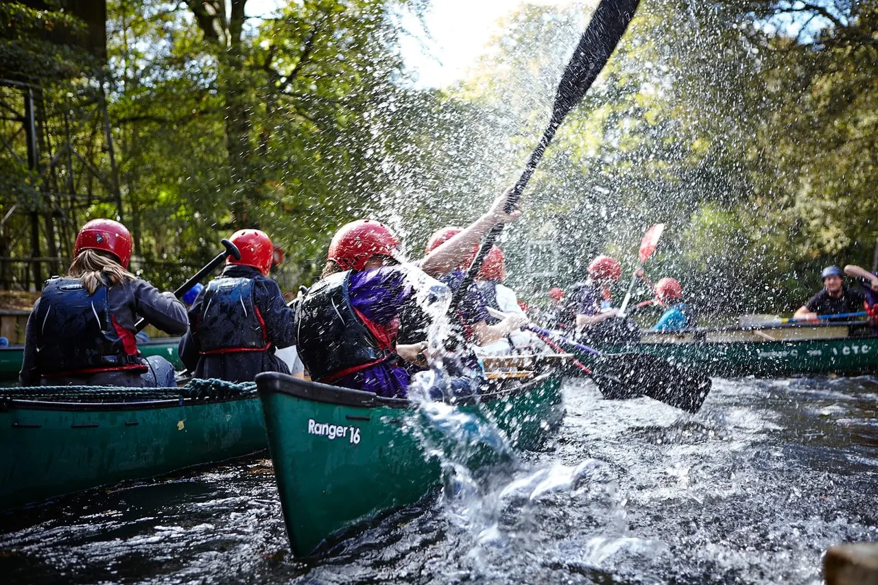 People in helmets and life jackets paddling canoes on a river with water splashing around them.