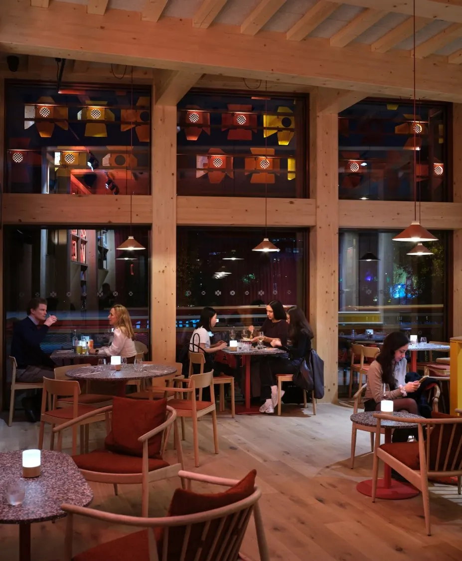 Modern wooden-beamed interior with diners seated at tables, large windows showing illuminated seating areas above, warm pendant lighting overhead.