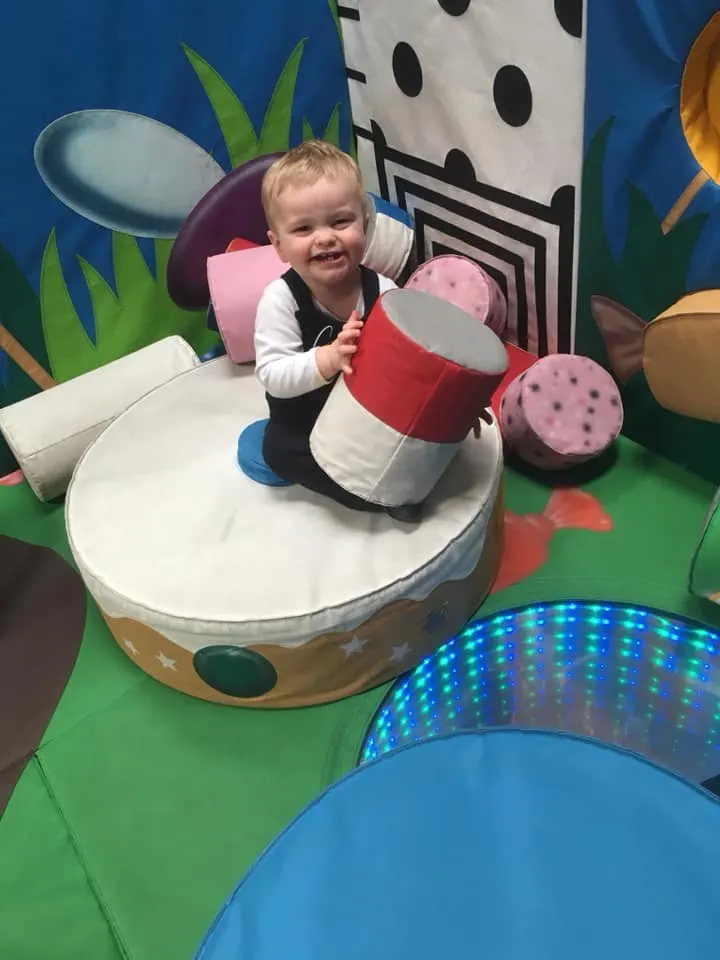 Toddler playing with oversized soft drum props in a colourful indoor play area with musical instrument decorations.