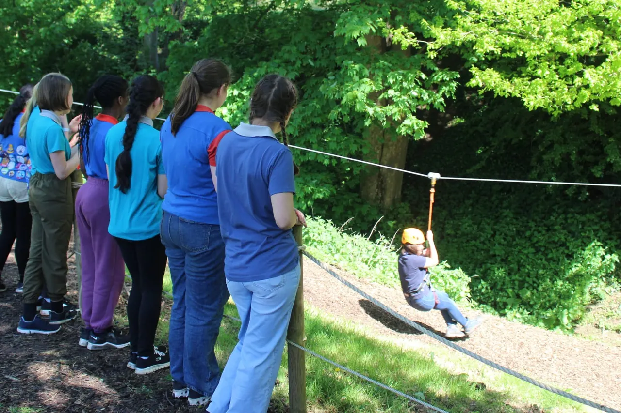 Group of children in blue uniforms watching a child zip-lining through a wooded outdoor adventure course.