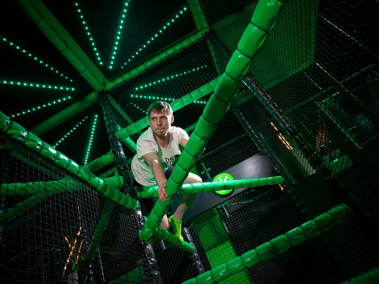 A man climbs through a neon-lit green foam obstacle course with illuminated tubes and mesh netting.