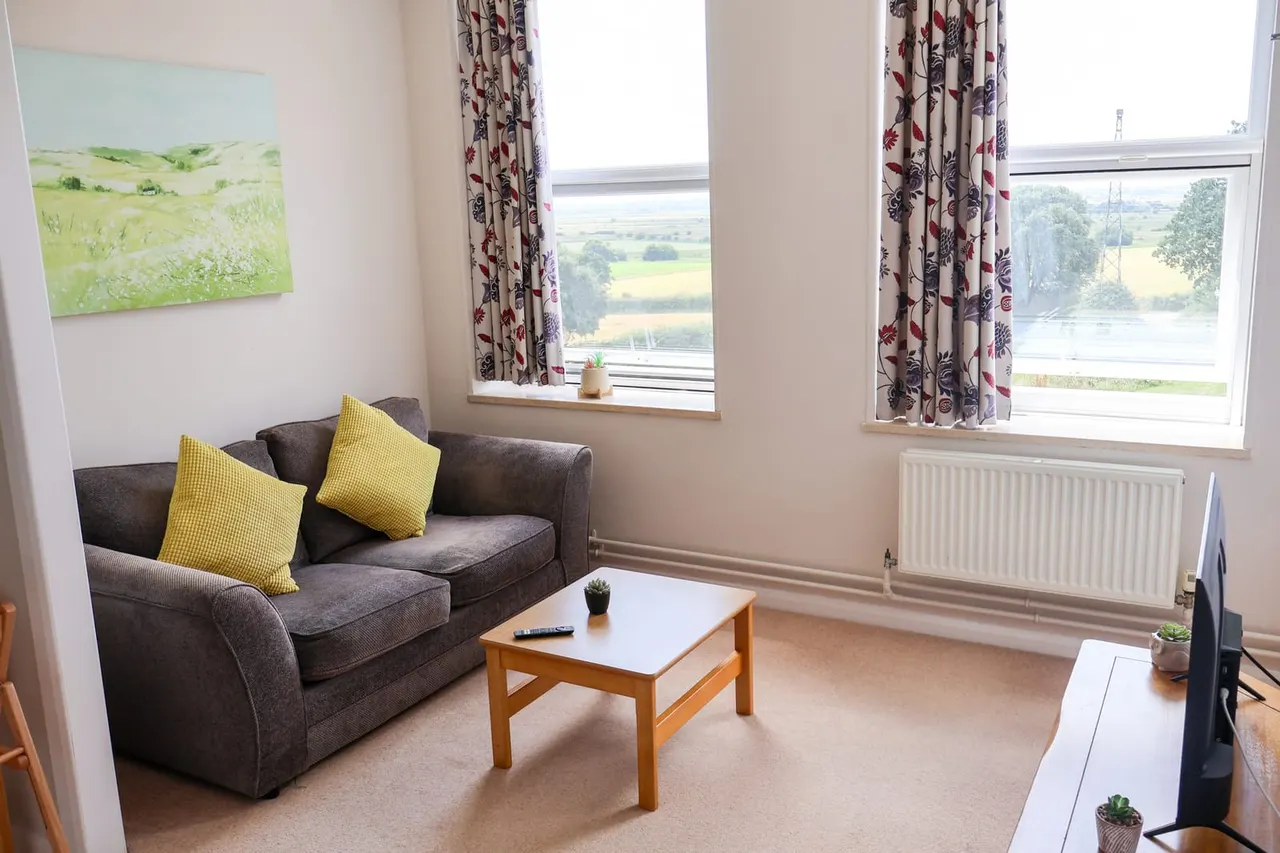 A bright, airy lounge with a grey sofa and yellow cushions, wooden coffee table, and large windows overlooking countryside and river views.