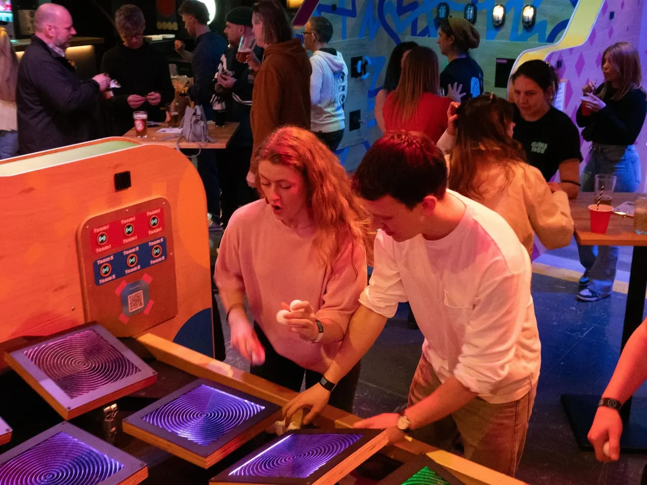 Two young people interact with illuminated interactive game controls at a colourful indoor entertainment venue whilst other visitors socialise in the background.