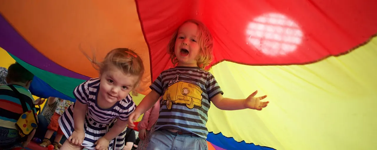 Two young children playing under a colourful rainbow parachute indoors.