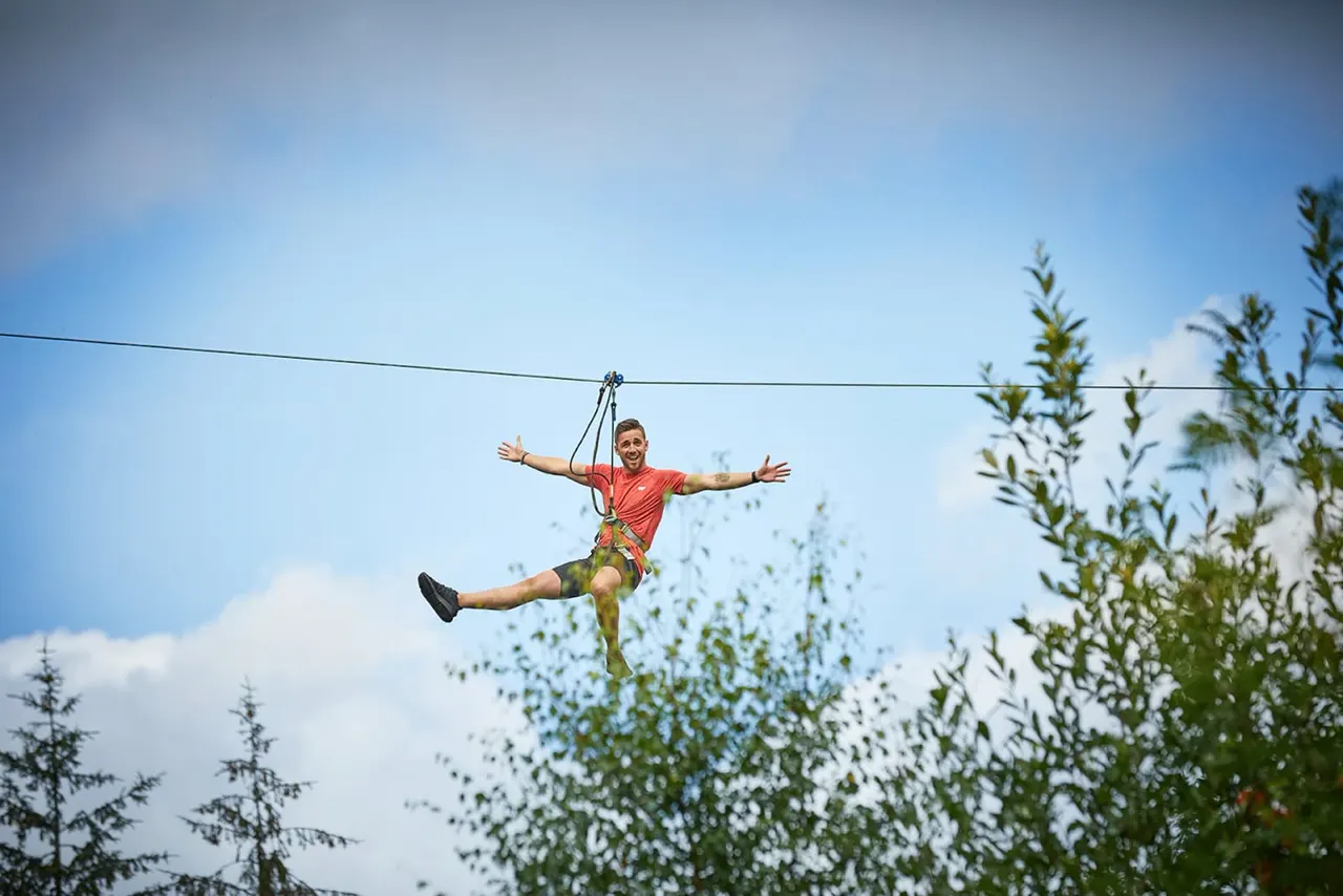 Person ziplining through a forest canopy with arms outstretched against a blue sky.