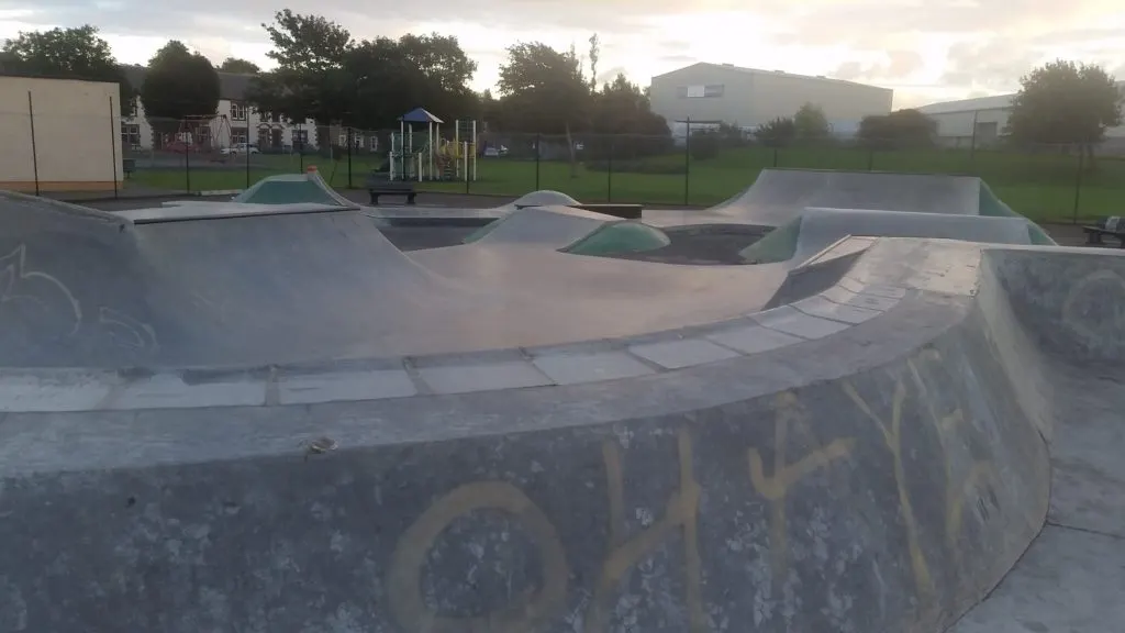 Concrete skate bowls and ramps at an outdoor skatepark with play equipment and green grass beyond.
