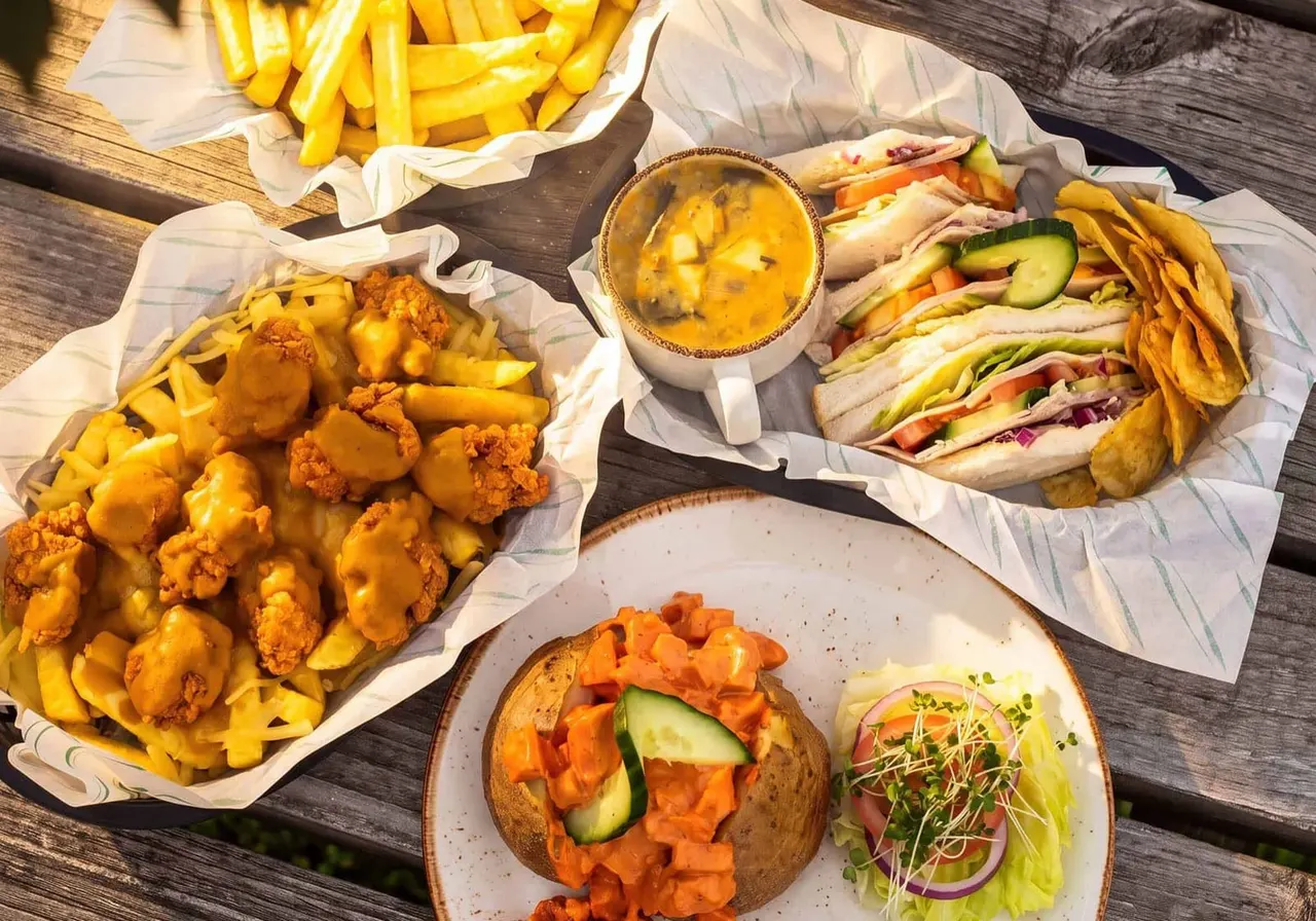 Overhead view of casual lunch spread with fried chicken and chips, sandwiches with vegetables, dipping sauce, and a loaded jacket potato on wooden table.