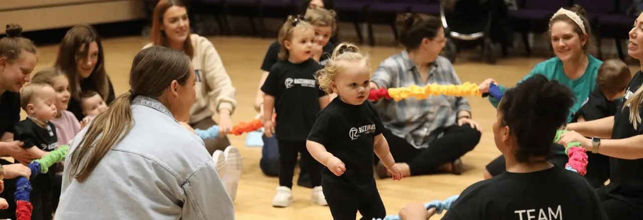 Toddlers and young children participate in a movement activity with adults in an indoor studio, holding coloured ropes.