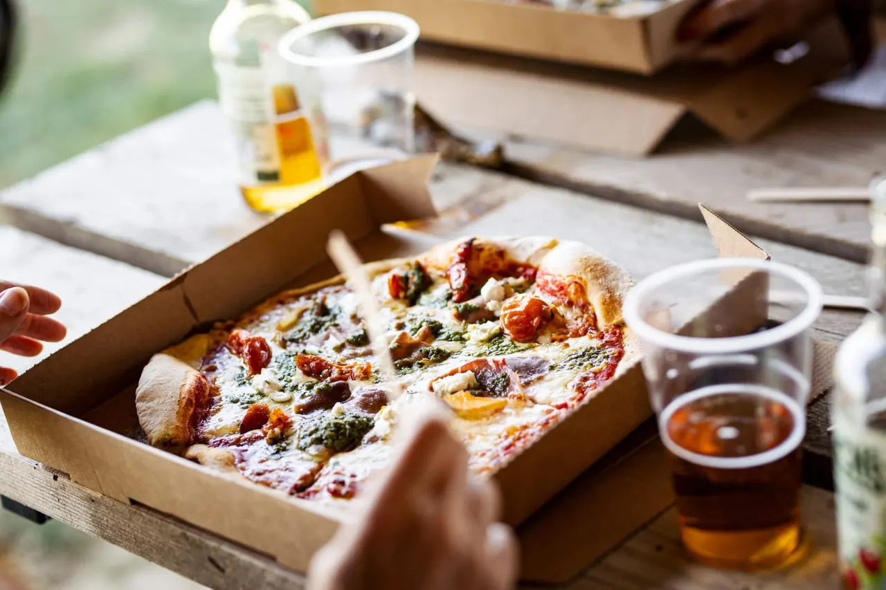 Pizza in a cardboard box on a wooden table with drinks and condiments, person's hand reaching for a slice.