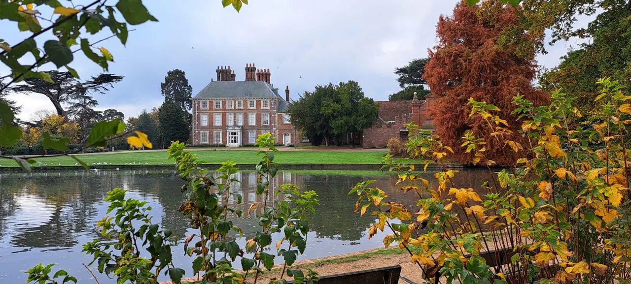 A red-brick Georgian mansion with manicured lawns reflected in a lake, surrounded by autumn-tinted trees and foliage in the foreground.