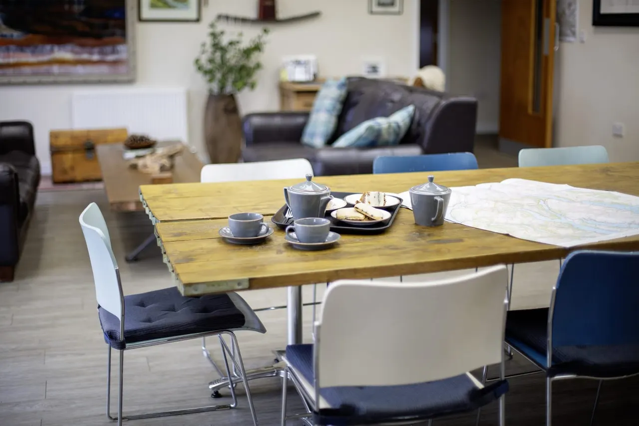 Wood dining table set with tea service, cups, saucers, teapot and biscuits, with modern chairs around it in a comfortable lounge area.