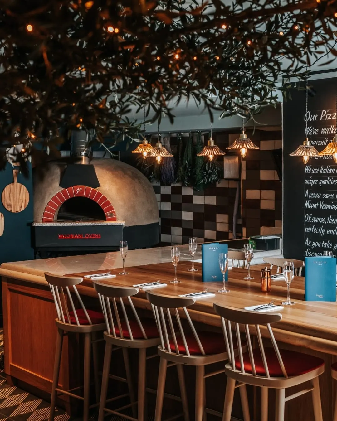 Dining counter with wooden chairs facing a wood-fired pizza oven, decorated with hanging lights and foliage.