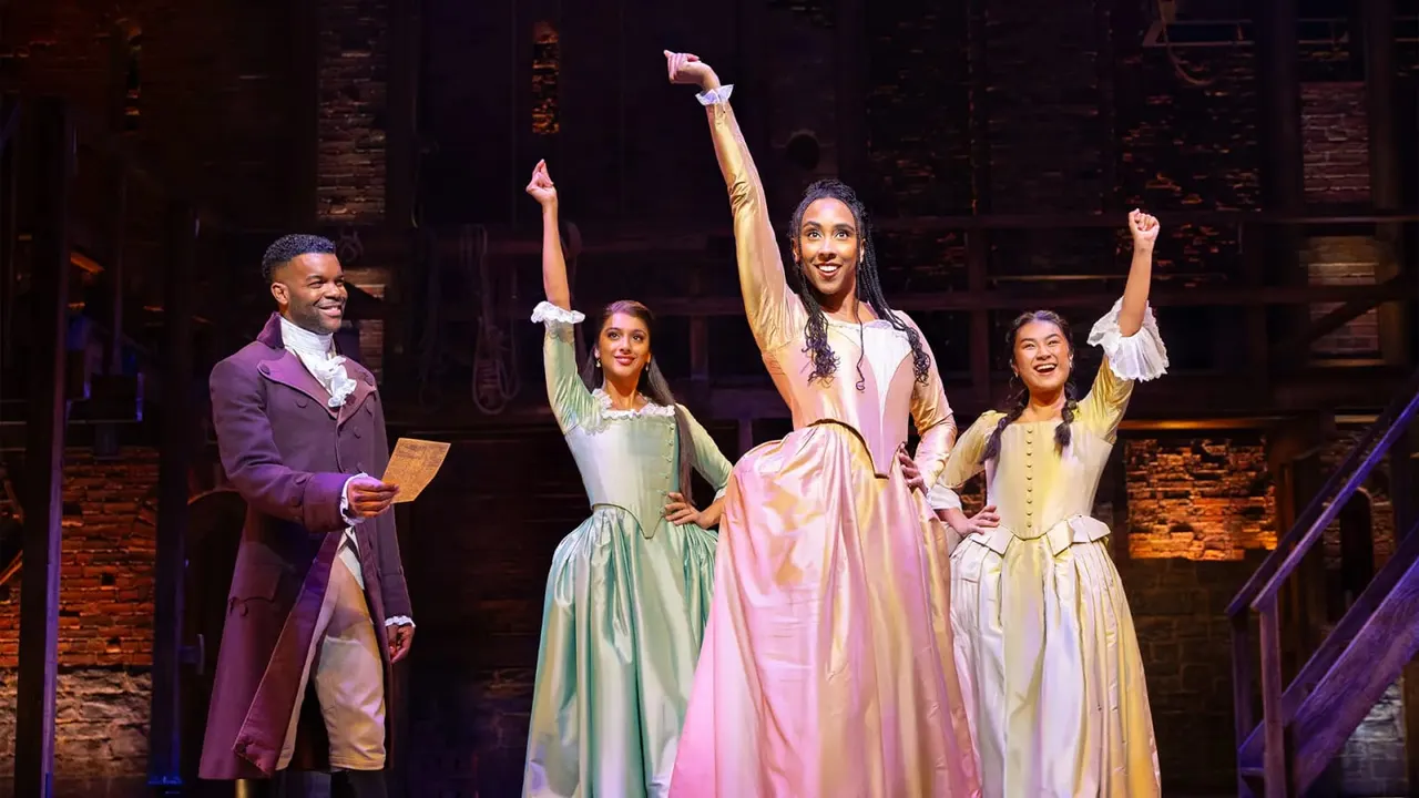 Cast members in period costume on stage during a theatrical performance, with raised fists in celebration.