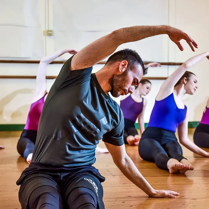 A man leads a yoga or fitness class, demonstrating a side stretch whilst seated on a wooden floor with other participants following along behind him.