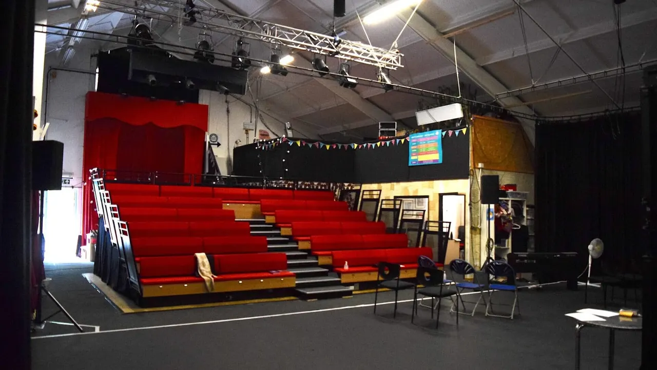 Indoor performance space with red tiered seating, stage with red curtain, theatrical lighting rig, and black backdrop with bunting.