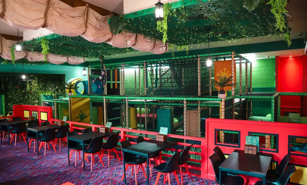 Colourful indoor soft play area with multi-level climbing structures, seating areas, and jungle-themed décor in green and red.