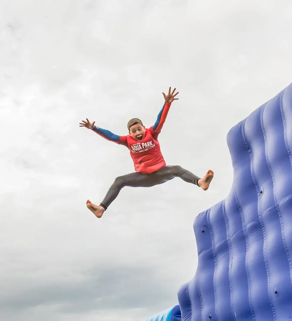 Child jumping off a large blue inflatable water slide with arms raised, against an overcast sky.