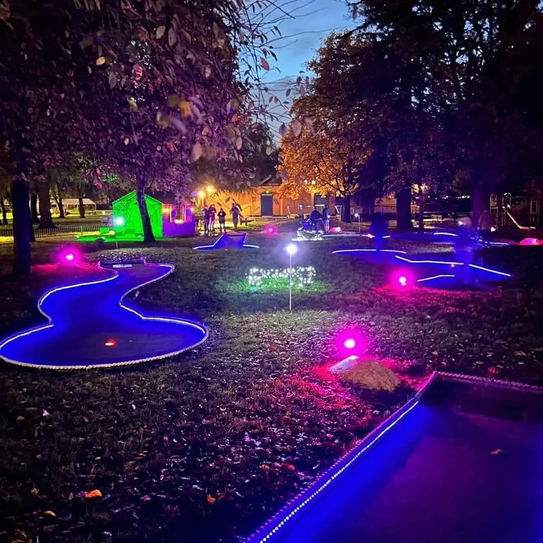 Night-time minigolf course with neon-lit blue and pink wavy paths, glowing obstacles, and a green clubhouse, trees lining the grounds.