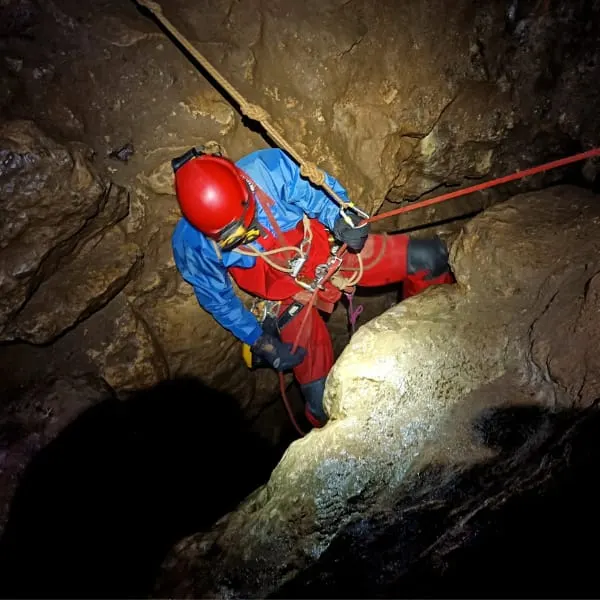 Person in blue and red gear abseiling down a rocky cave wall with safety equipment.