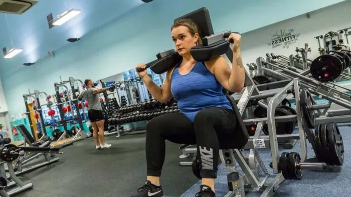 Woman exercising with barbell in a modern gym with equipment and other members visible.