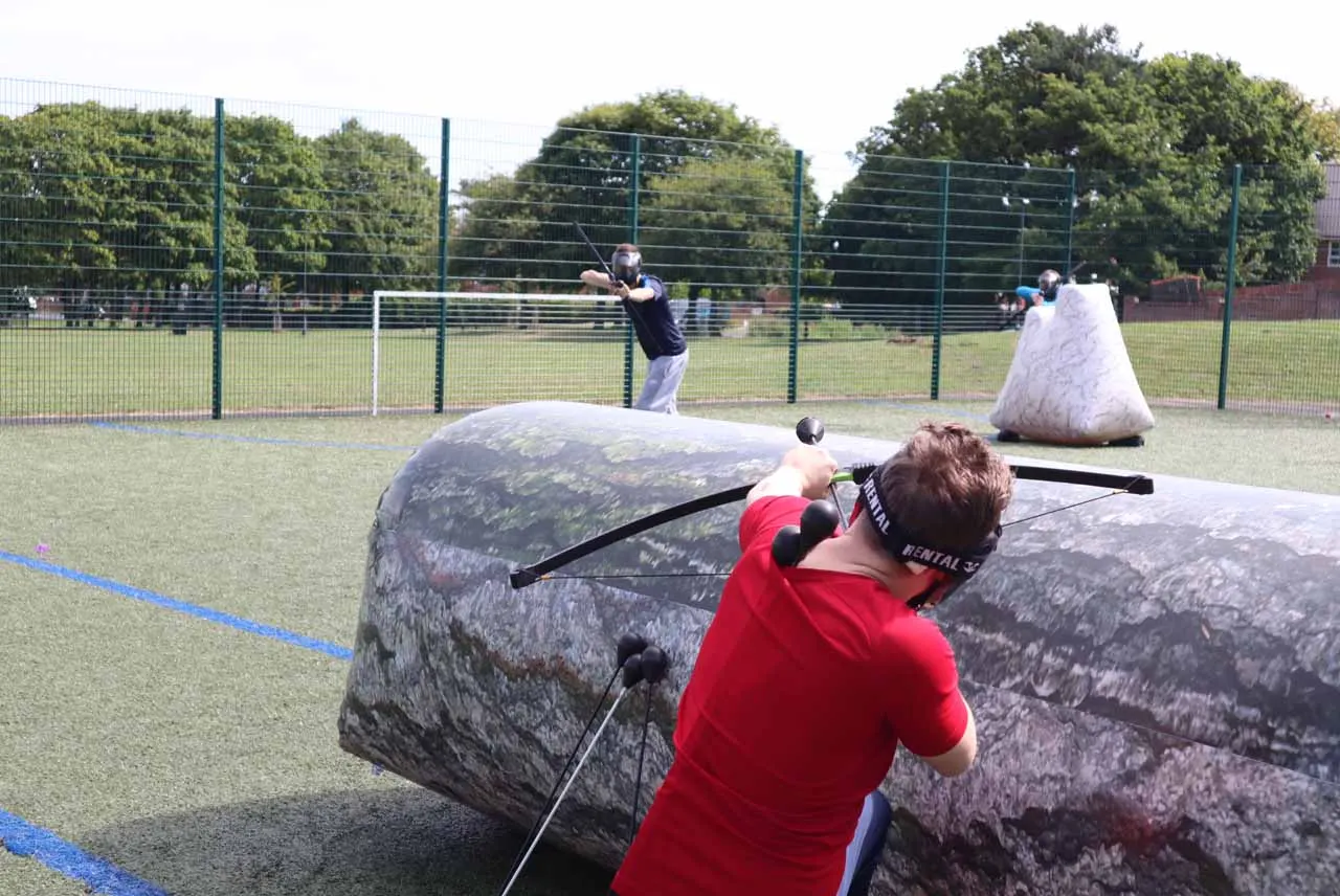 Young people playing bubble football in an enclosed outdoor sports arena, with inflatable bumpers and fenced pitch.