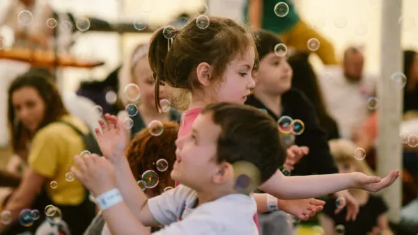 Children playing with soap bubbles indoors with adults watching in the background.