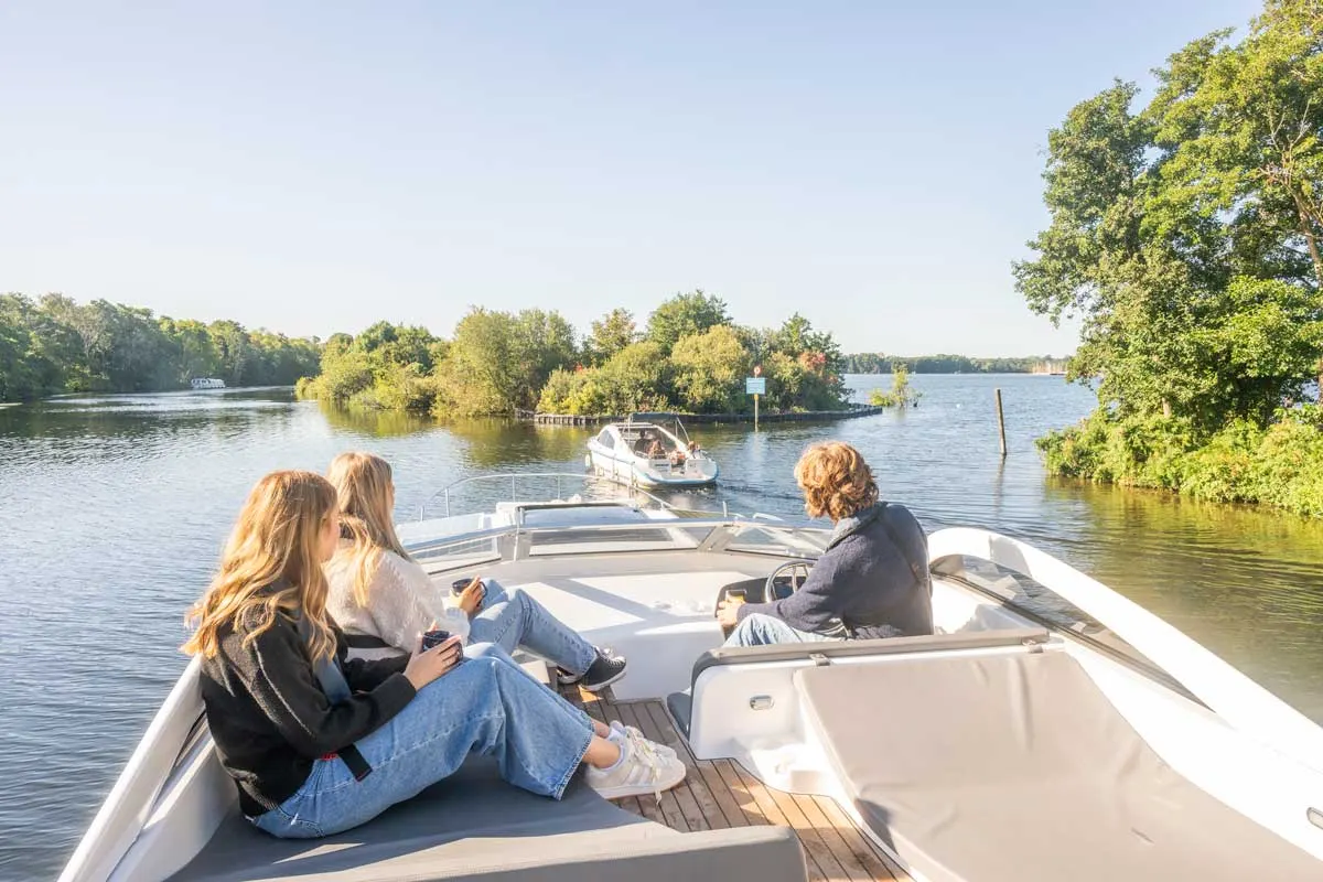 Three people cruising on a motorboat along a tree-lined river on a sunny day.