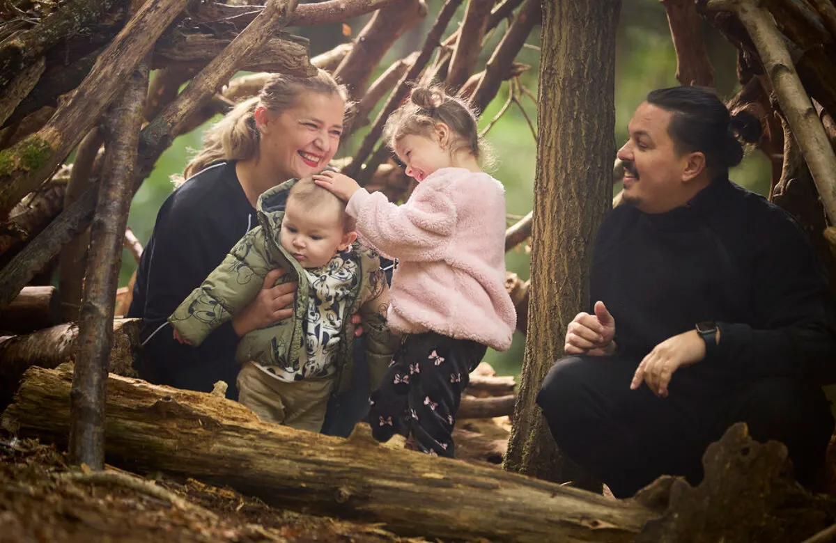 Family with two young children playing inside a wooden den made from branches and logs.