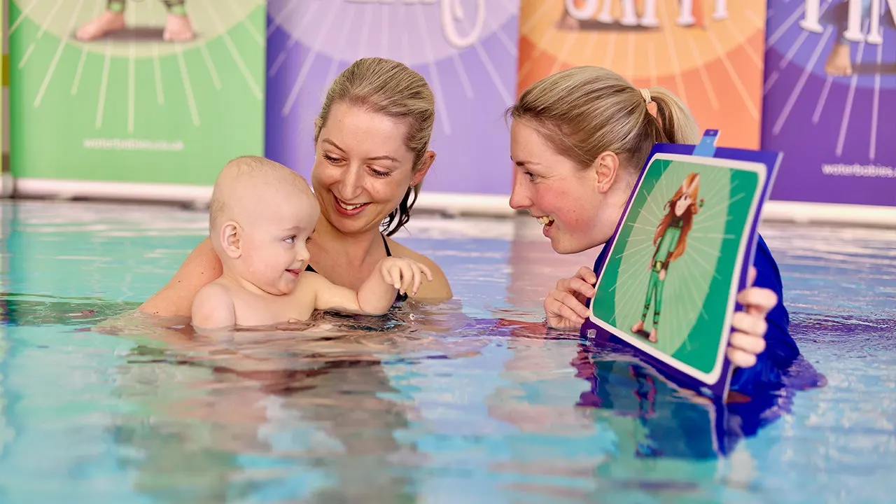 Two instructors and a toddler in a swimming pool during a lesson, with one instructor holding a visual aid card.