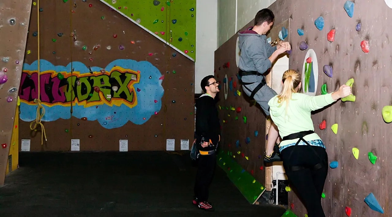 People climbing an indoor rock wall with colourful handholds whilst wearing harnesses.