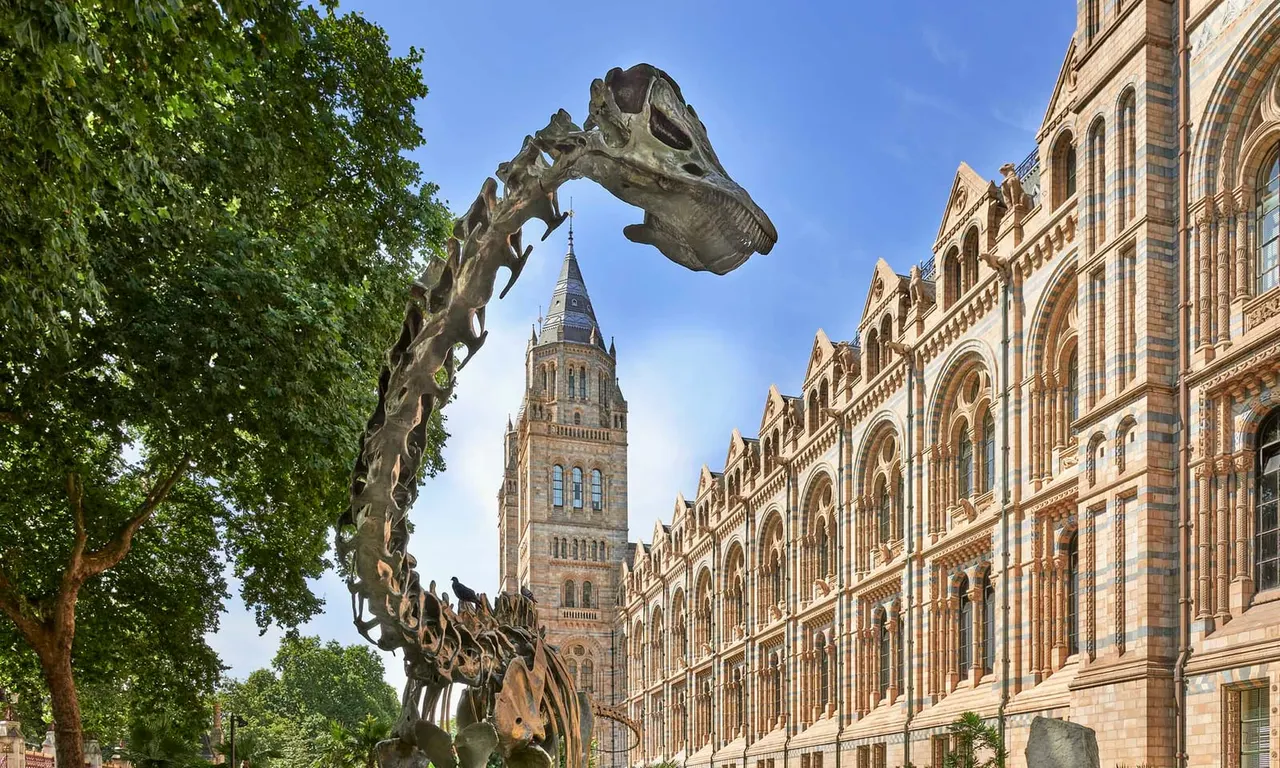 A dinosaur skeleton sculpture towers above a tree-lined street facing the Natural History Museum's Victorian Gothic building and clock tower.
