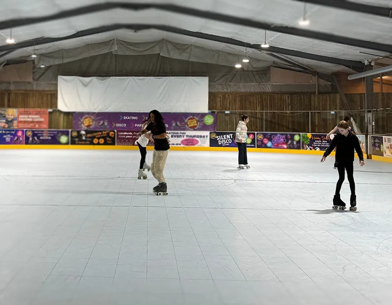 Several people roller skating on an indoor rink with sponsor boards and a projection screen.