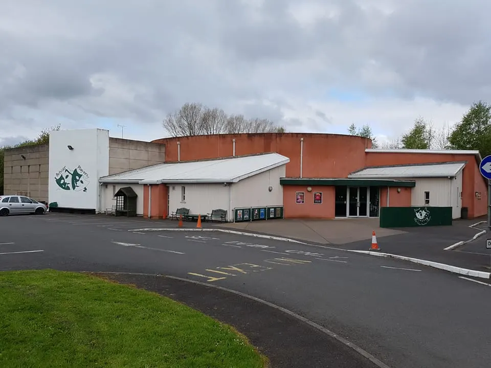 Exterior view of a modern leisure venue with red-brick and white rendered walls, multiple entrances, parking area and grass verge on an overcast day.