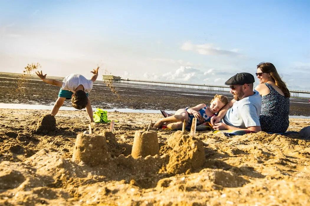 Family playing and building sandcastles on a sandy beach with a pier visible in the background.
