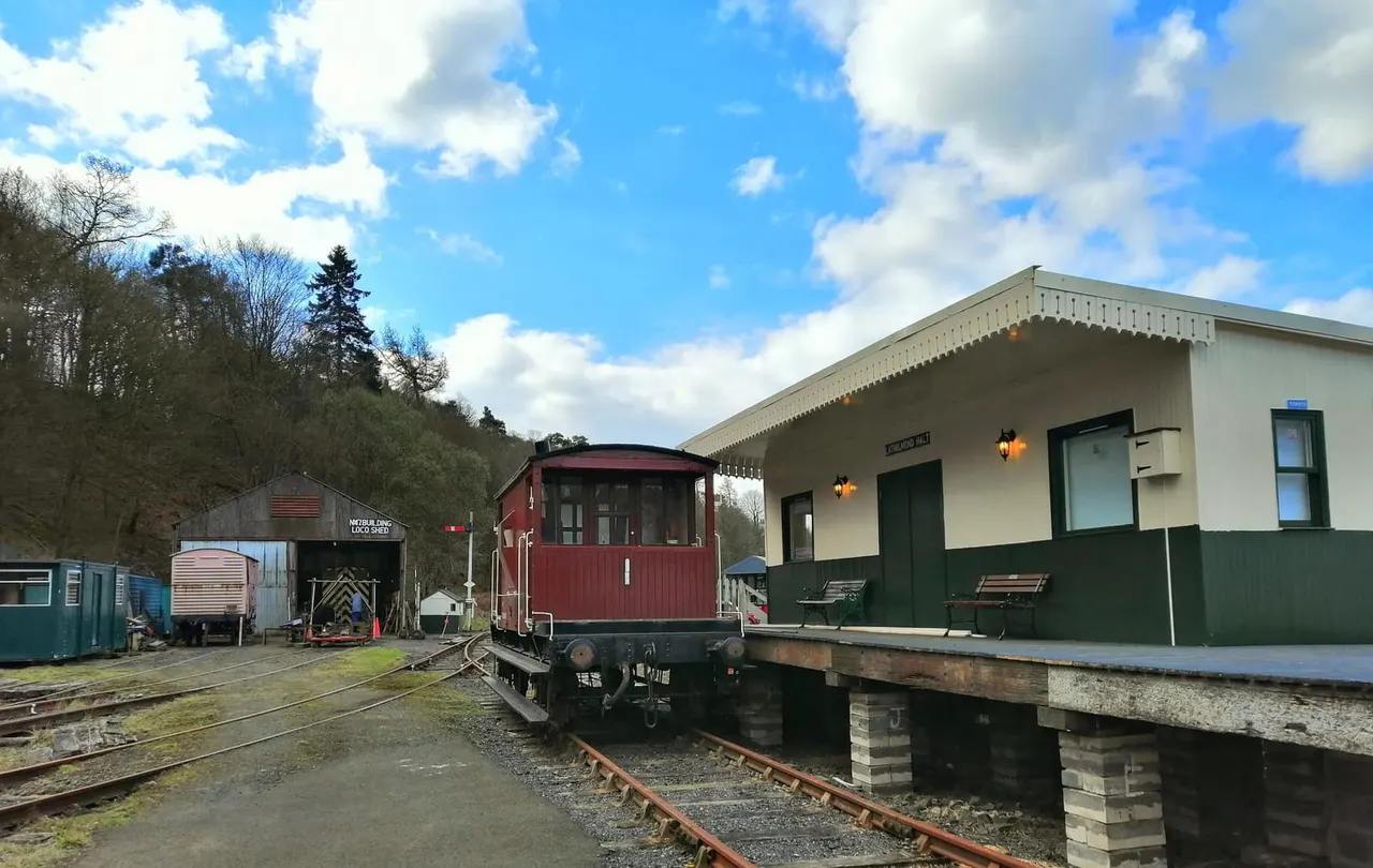 Heritage railway depot with a red vintage carriage on tracks, stone station building, and outbuildings against woodland backdrop.