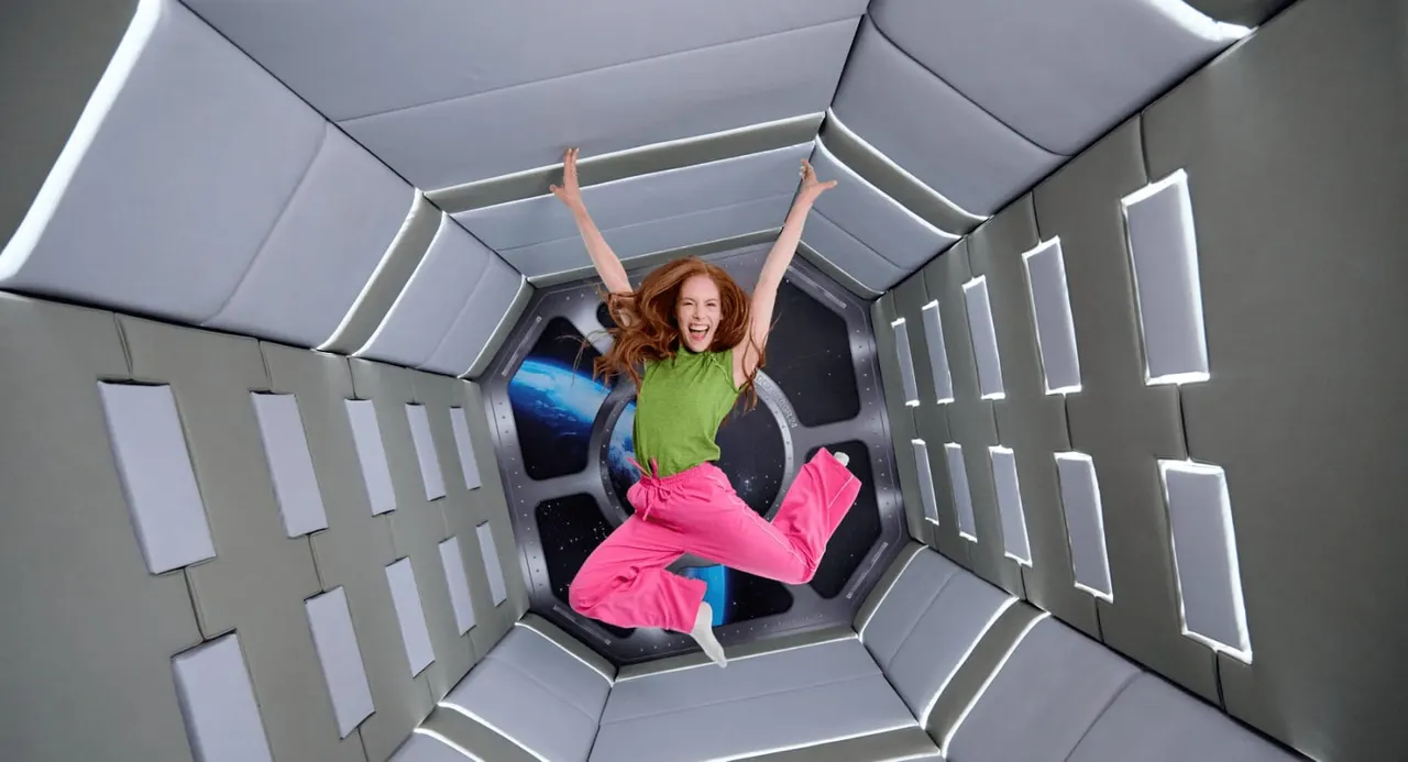Woman floating in zero gravity inside a spacecraft simulator chamber with arms raised in joy.