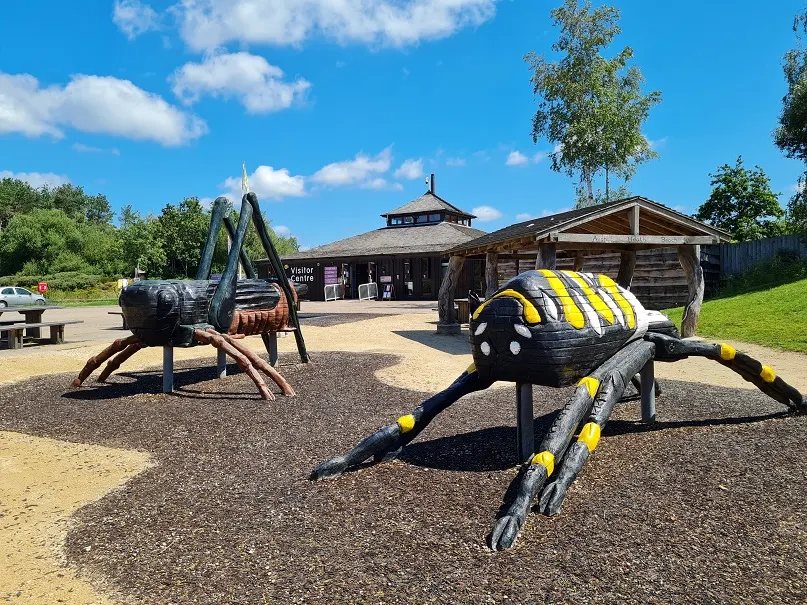 Giant insect-shaped climbing structures in a playground with wooden visitor centre buildings and green fields under blue sky.