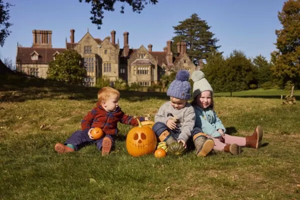 Three young children sit on a lawn with carved pumpkins in front of a large brick manor house.