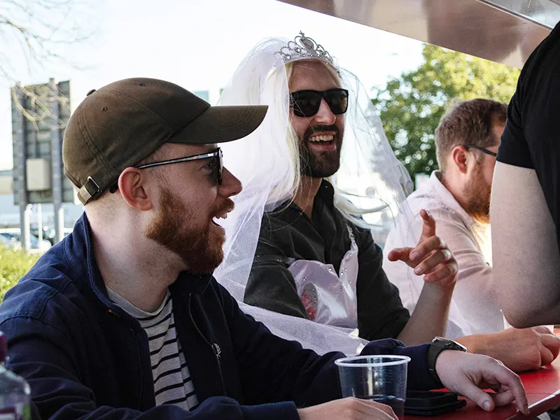 Three men laughing together on an outdoor seated activity, one wearing a white veil and tiara, in bright sunshine.
