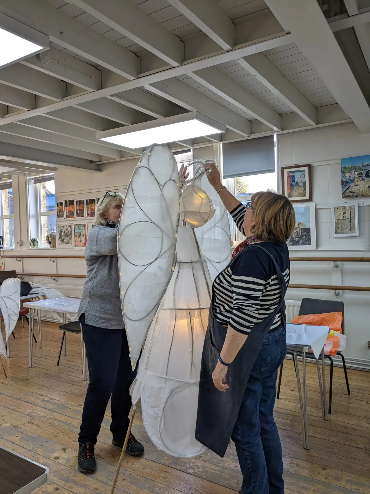 Two women assembling a large white fabric art installation with black line patterns in a bright studio space.