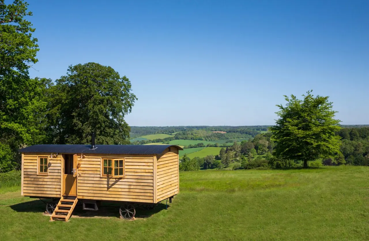 Wooden shepherd's hut on wheels in a meadow with rolling countryside and tree-lined hills beyond.