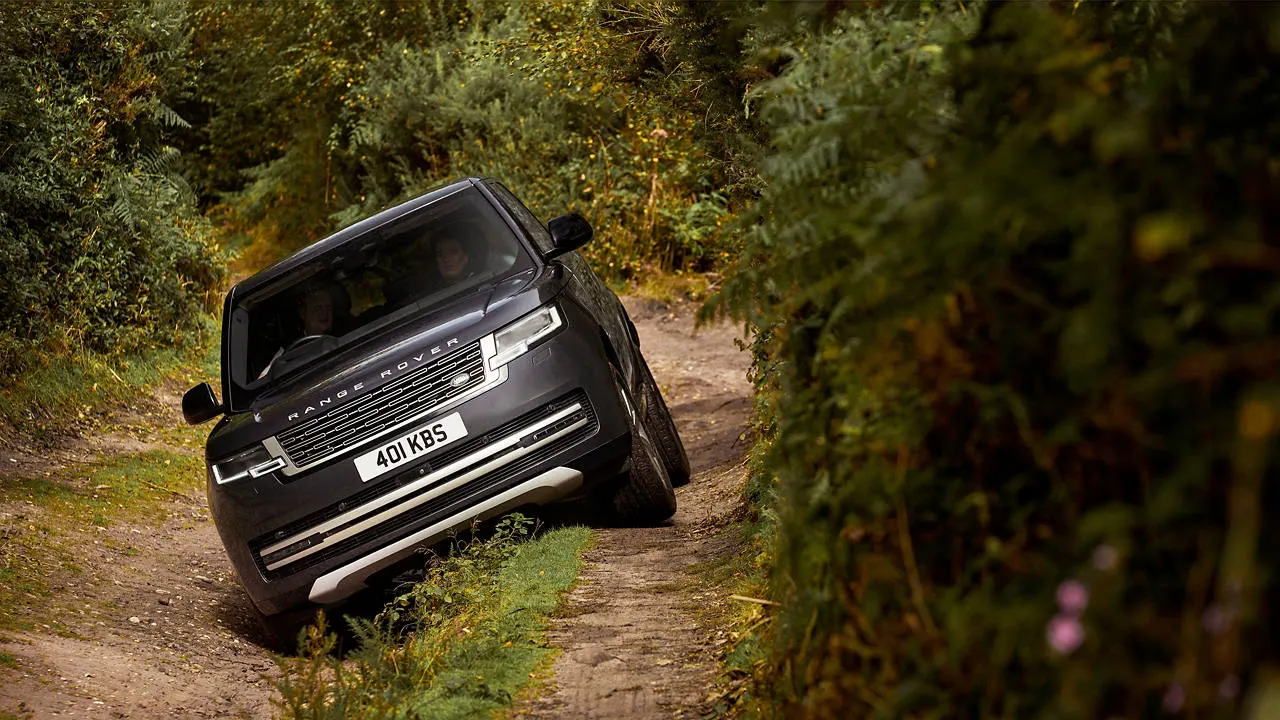 A black Range Rover drives along a narrow, tree-lined dirt track through woodland.