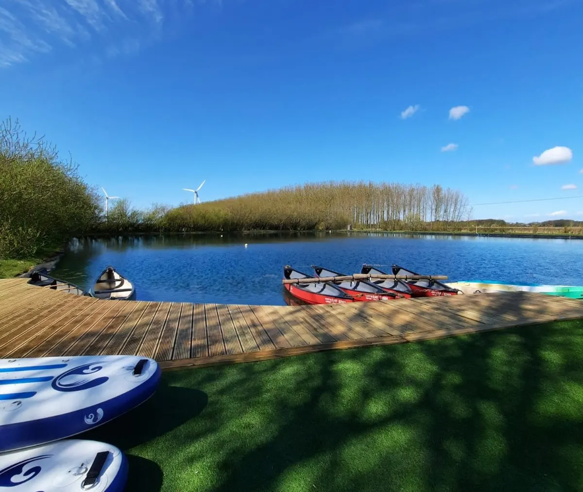 Wooden deck beside a calm blue lake with kayaks and paddleboards lined up, reed-lined banks and wind turbines visible in the distance.