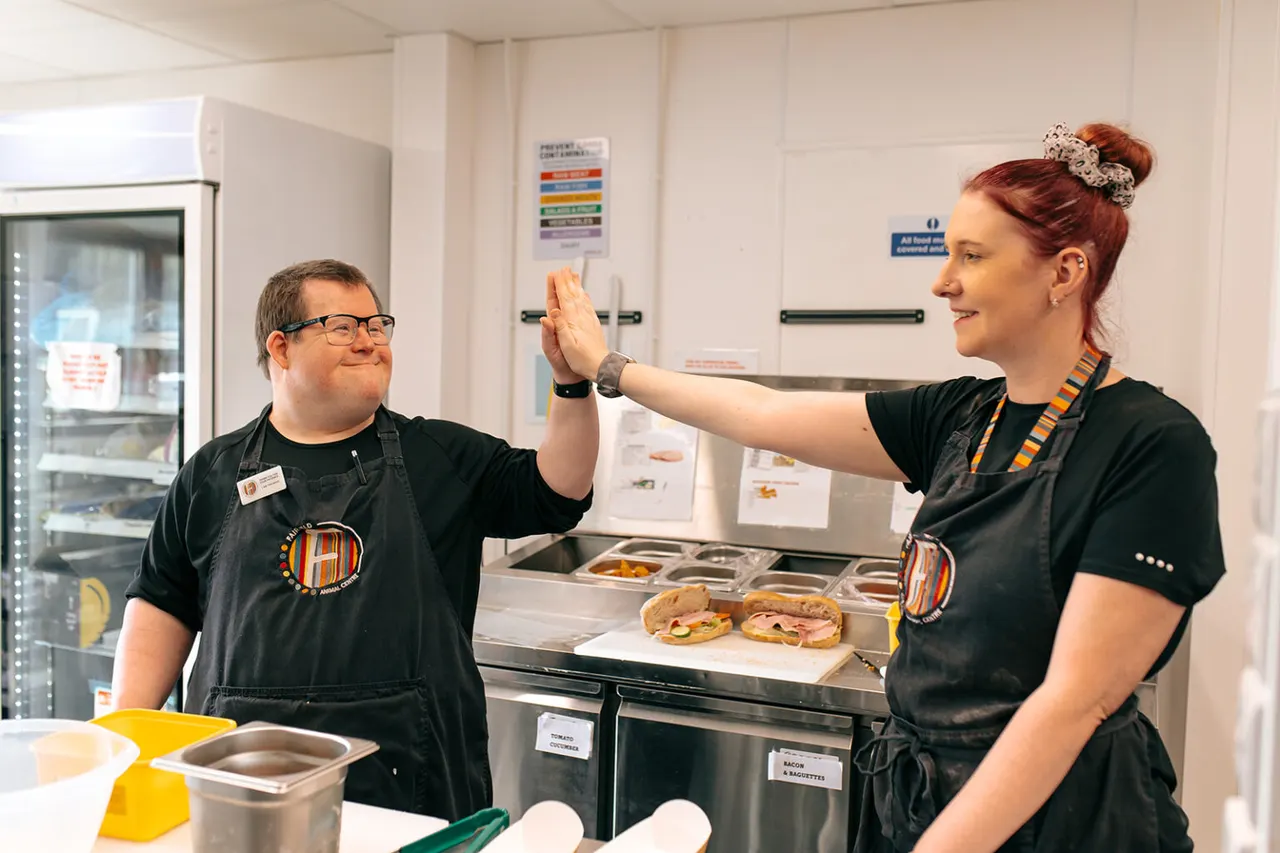 Two staff members give each other a high five in a kitchen, with sandwiches and food preparation equipment visible.