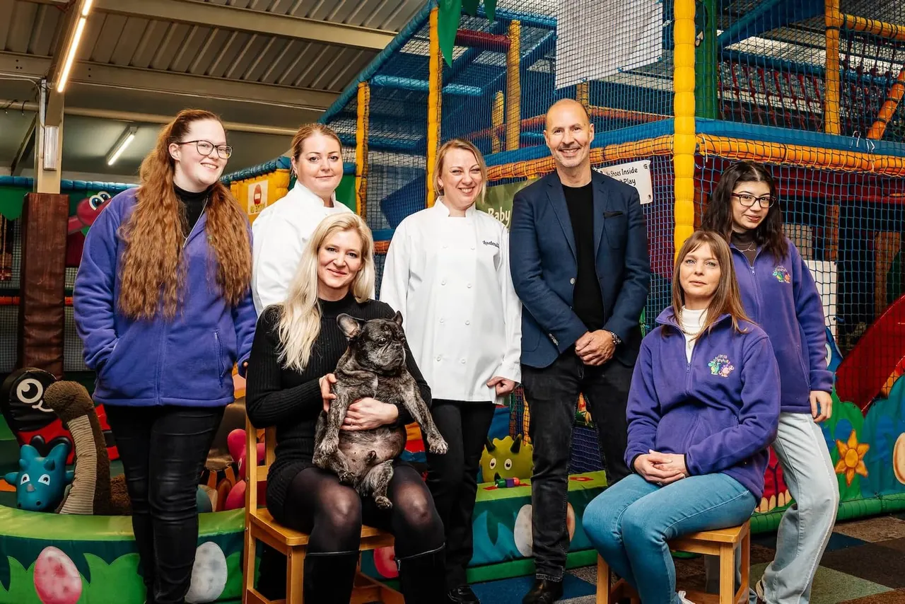 Seven adults and a French bulldog pose together indoors at a colourful soft play facility with climbing structures.