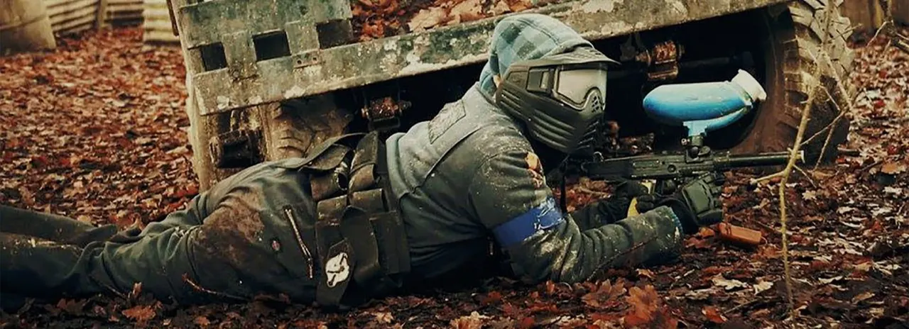 Player in protective gear and mask crouching behind cover whilst aiming paintball marker during game.