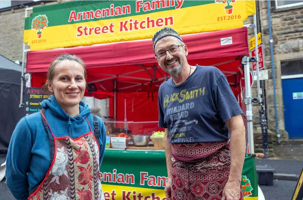 Two people stand smiling in front of a brightly coloured Armenian Family Street Kitchen food stall.